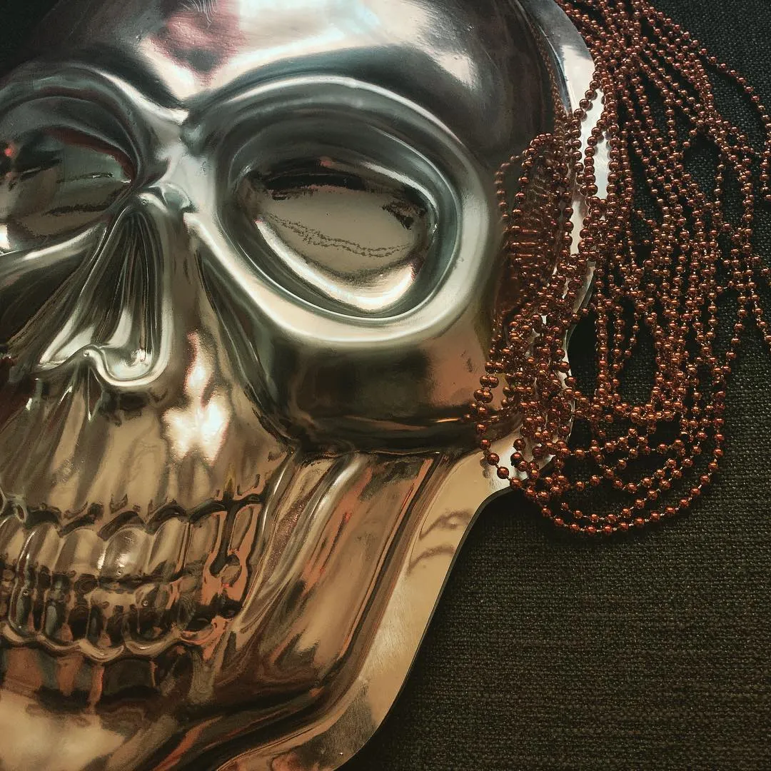 A moody promo teaser for Hearing Pox: a shiny silver skull mask draped with strands of copper-colored beads, lit against a black background.