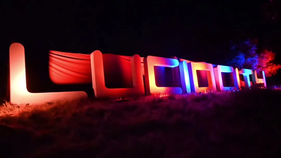 The giant illuminated LUCIDITY letter sculpture glowing in rainbow colors — reds, oranges, blues, and purples — on a dark festival field at night.