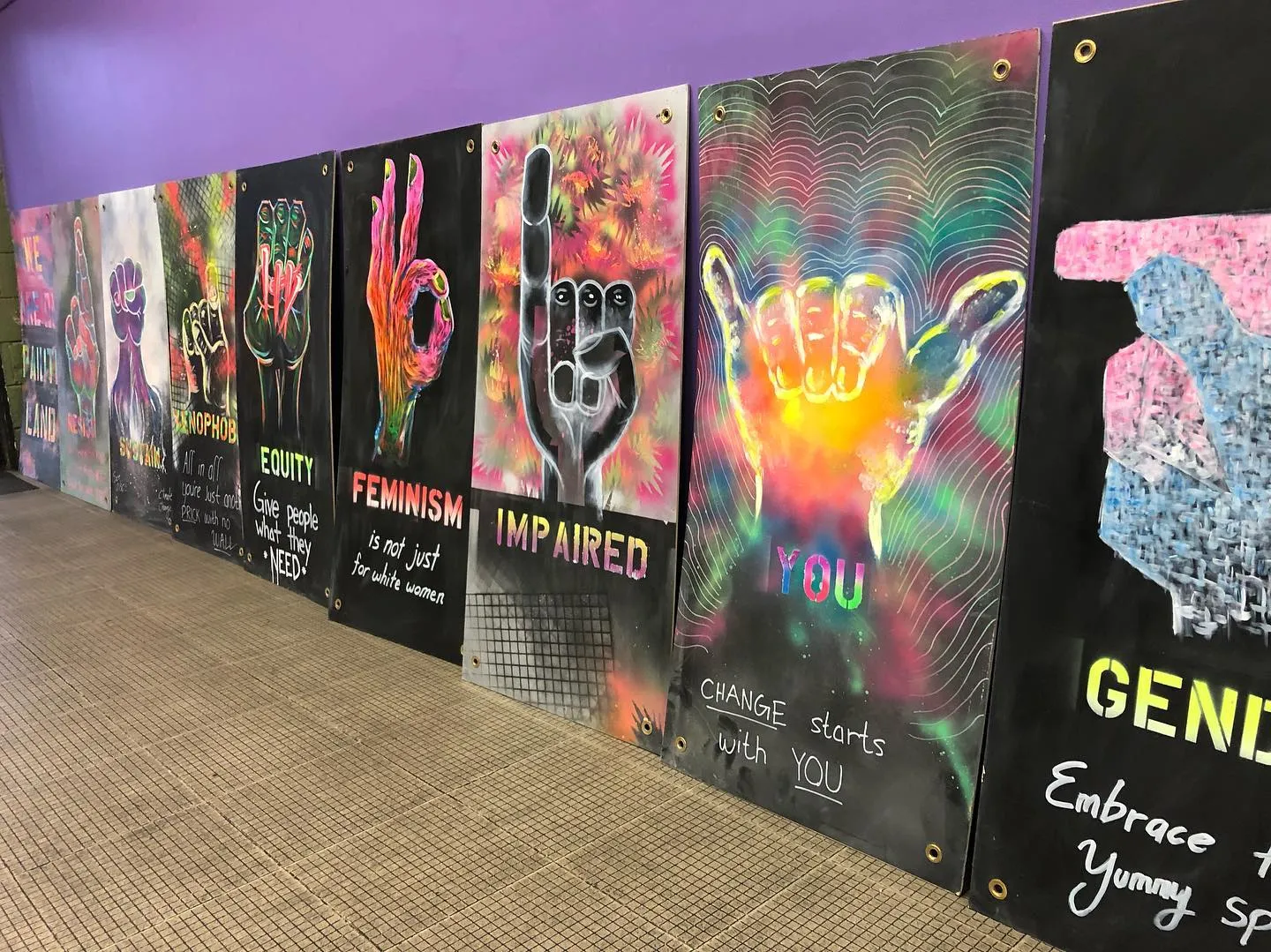 A row of ASL Love's psychedelic blacklight art panels leaned against a purple wall at the Deaf Community Center. Each tall panel shows a glowing neon ASL handshape paired with a word painted in vivid pinks, greens, and blues on black — among them FEMINISM ('is not just for white women'), IMPAIRED, EQUITY ('Give people what they NEED'), and YOU ('Change starts with YOU').