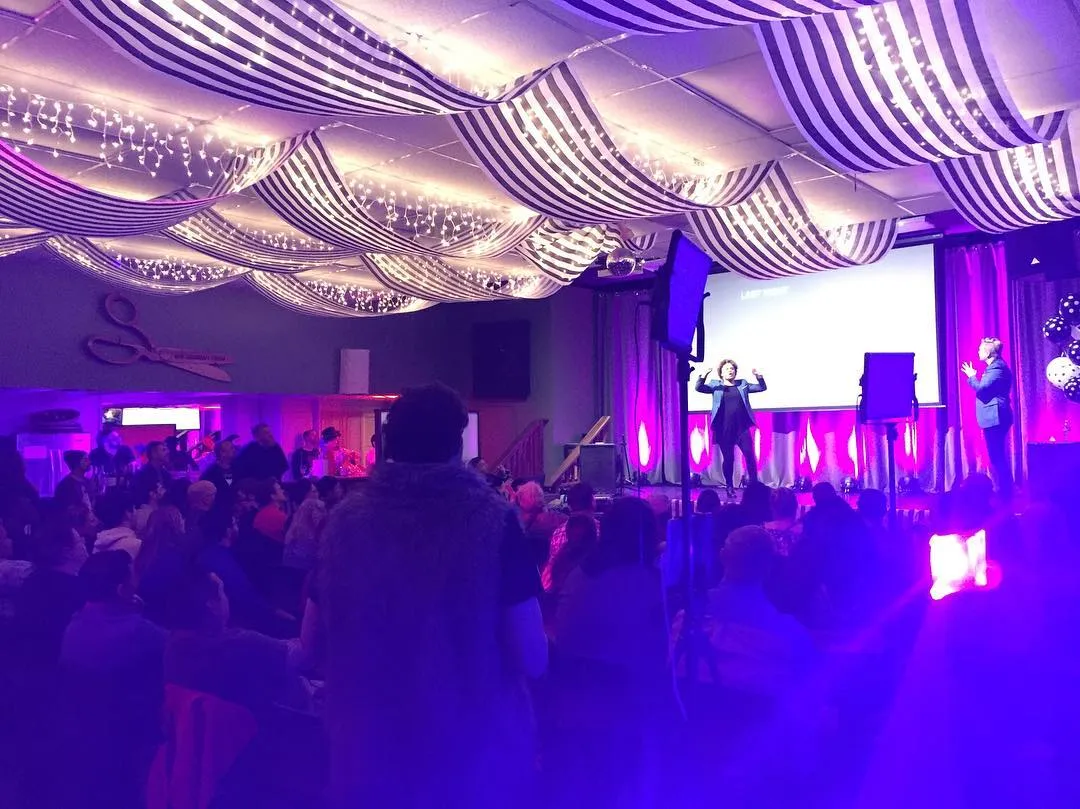 Festival of Fools in the Deaf Community Center hall, bathed in purple light beneath a draped, fairy-lit ceiling. A performer stands center stage signing to a packed, seated audience.