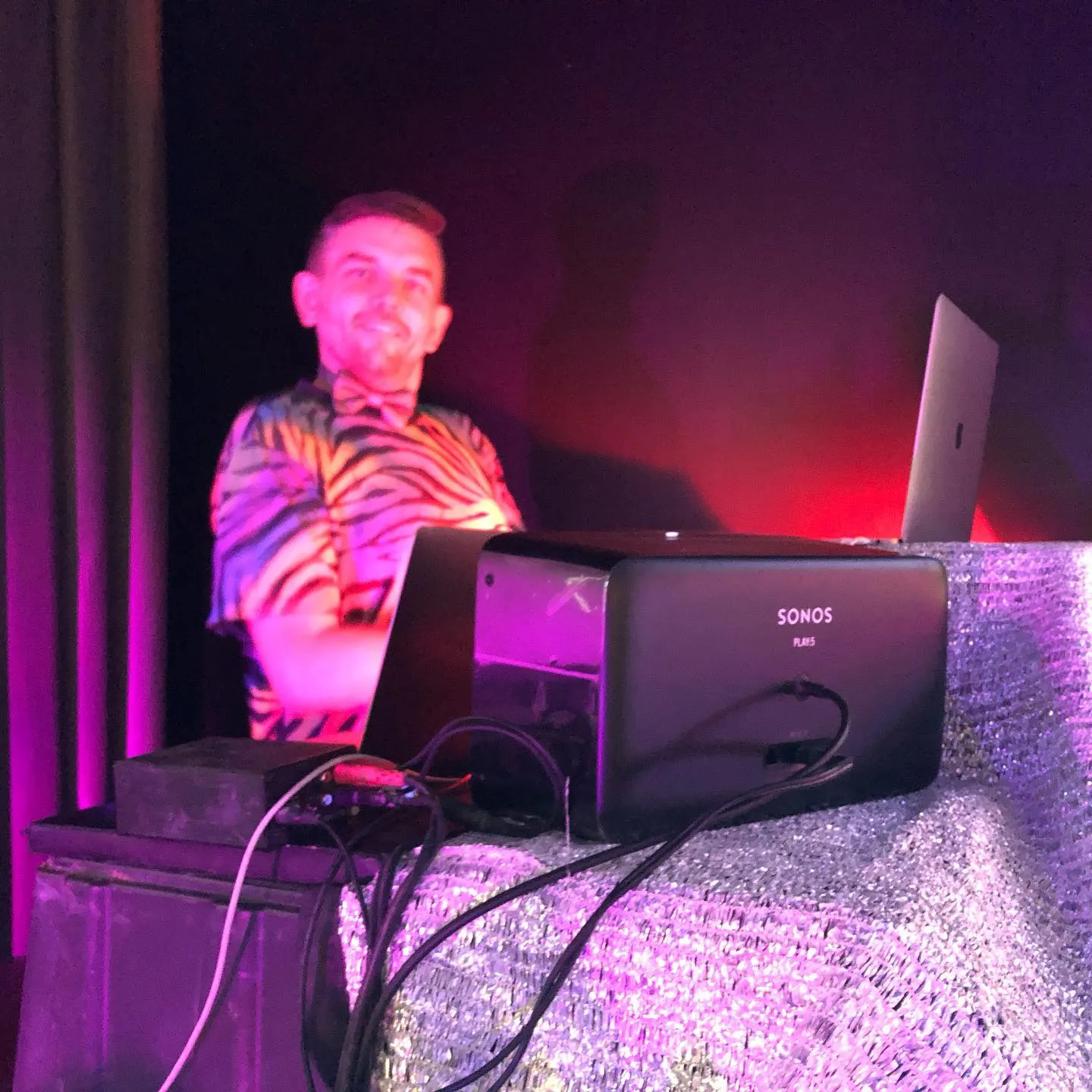 A DJ in a tiger-print shirt and bow tie grins behind the decks at Fire and Vice — a laptop and a Sonos speaker on a silver sequined table, lit in hot pink and red.