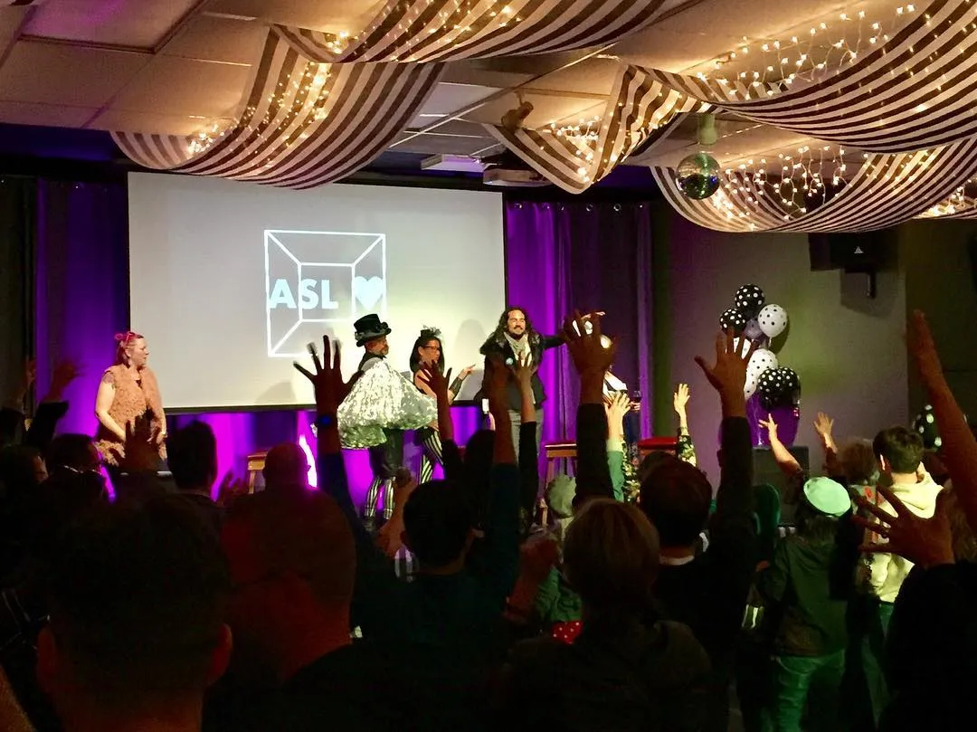 The Festival of Fools cast on stage in front of a projected ASL Love cube-and-heart logo, while the audience raises their hands to sign applause. Balloons and string lights frame the room.