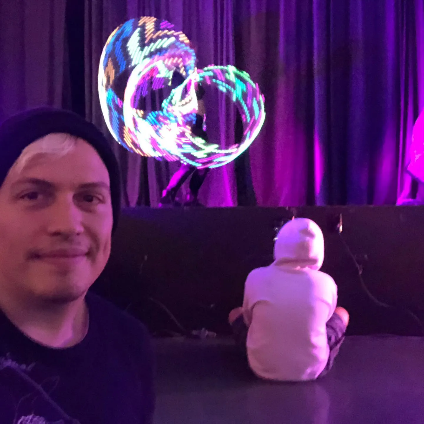 An LED hoop performer spins glowing hoops into bright looping trails of blue, pink, and green on the Fire and Vice stage, watched by a child seated on the floor in a white hoodie.