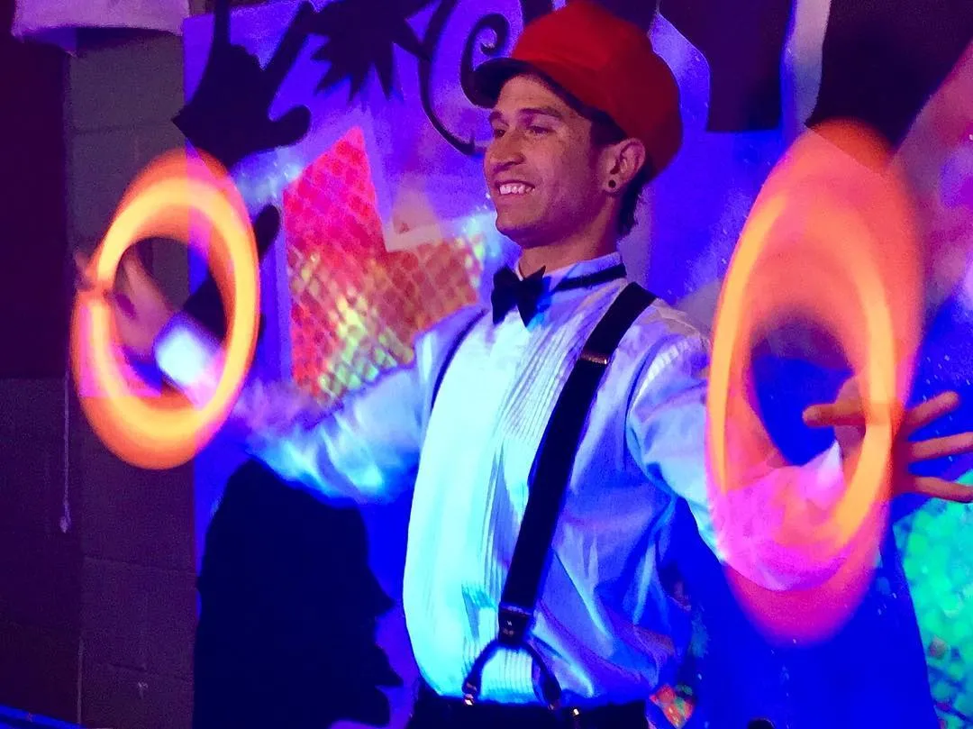 A Festival of Fools flow-arts performer in a red cap, bow tie, and suspenders smiles as he spins glowing orange LED poi, their light trails arcing on either side against ASL Love's art backdrop.